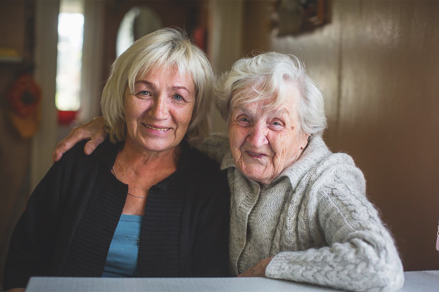 Ältere Frau mit einer jüngeren Angehörigen, Arm in Arm zu Hause — würdevolle Betreuung im vertrauten Umfeld